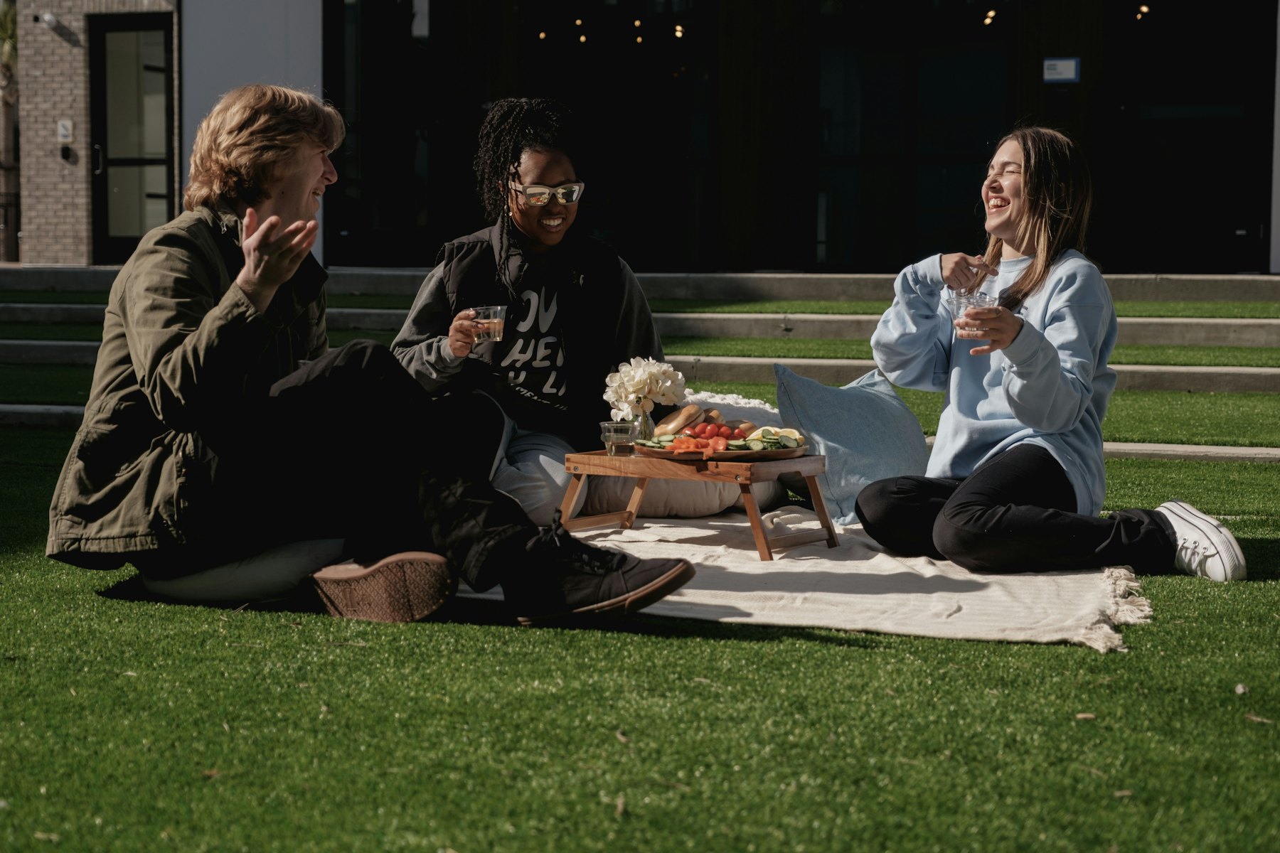 Three friends laughing on a blanket on a sunny lawn