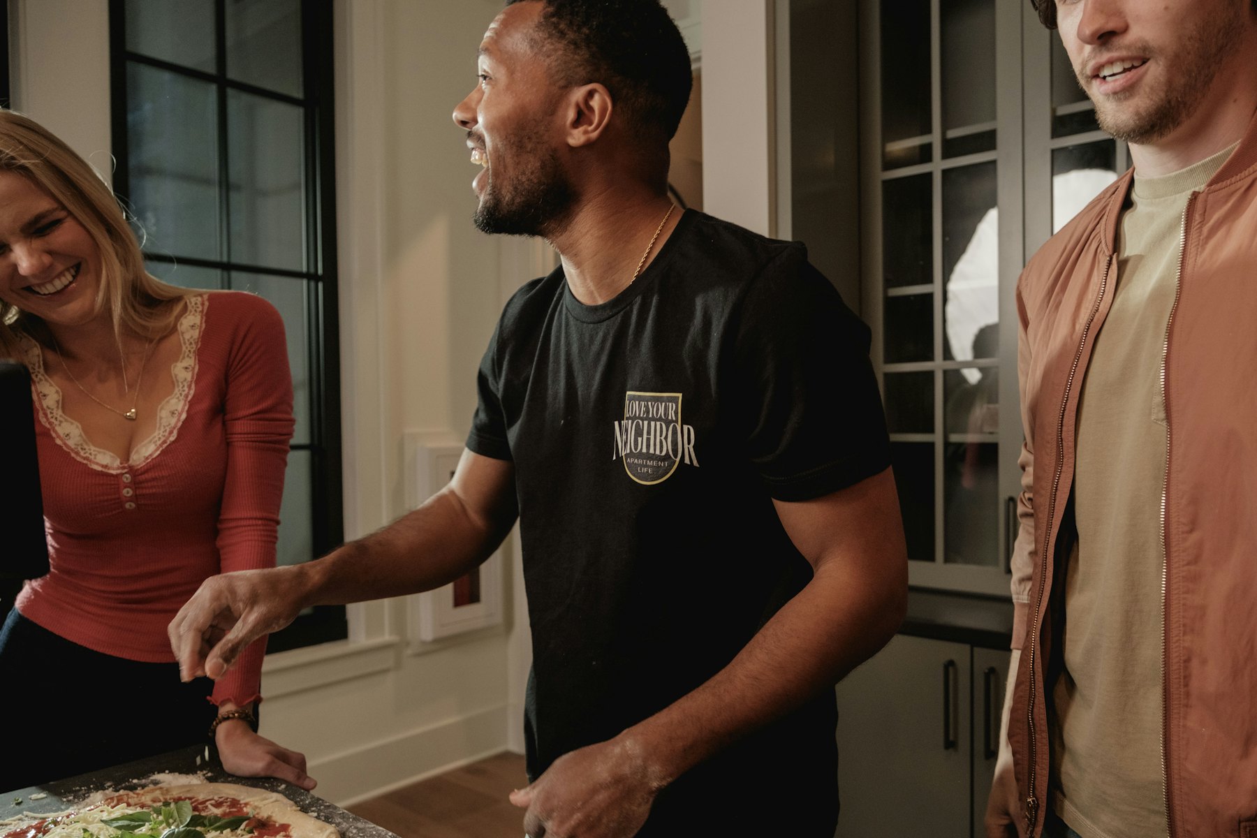 Three friends laughing while making pizza in a kitchen