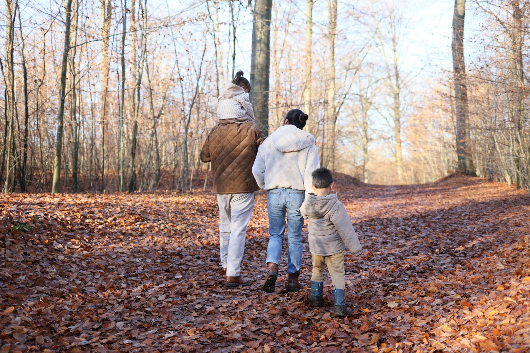 A family walking away down a path covered in autumn leaves, a child on one parent’s shoulders